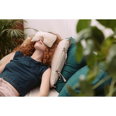 Kobieta z rudymi kręconymi włosami leży na jasnej poduszce w otoczeniu zielonych poduszek i roślin, z lawendową poduszką na oczy do relaksacji.